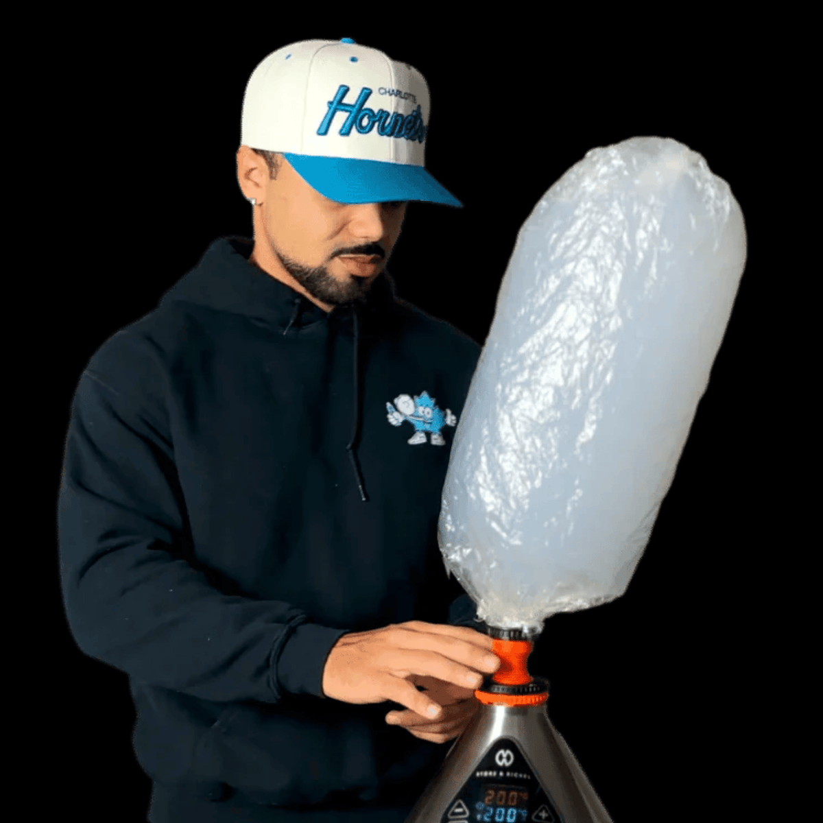 Classic Volcano Storz & Bickel - Man wearing a black hoodie and blue cap attaches a clear balloon to a black-and-orange vaporizer device.